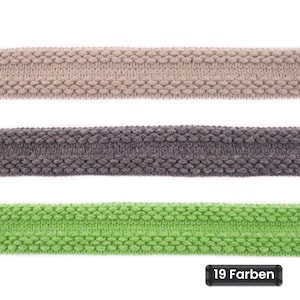 Op de afbeelding: Drie horizontale stroken gebreid lint, elk met een gestructureerd nopjespatroon langs de randen en een glad gebreid midden. De stroken zijn beige, donkergrijs en felgroen van kleur. Een zwart label toont de tekst 19 Farben.