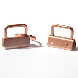 Può includere: Due clip per ferramenta in metallo color oro rosa con una parte superiore ad anello rettangolare e una base piatta e larga. Una clip presenta un anello diviso circolare attaccato. La ferramenta ha una finitura metallica liscia e una barra a U.
