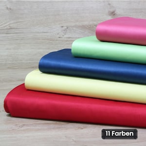 Könnte beinhalten: Ein Stapel aus fünf gefalteten Stoffballen in den Unifarben Rot, Hellgelb, Dunkelblau, Hellgrün und Magenta. Der Stoff hat eine glatte, satinartige Oberfläche. Ein schwarzes rechteckiges Etikett in der Ecke zeigt den Text 11 Farben.