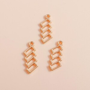 Puede incluir: Tres dijes de metal en tono dorado, cada uno con una disposición vertical de cuatro marcos rectangulares abiertos conectados. Cada dije incluye un pequeño lazo circular en la parte superior para su fijación, con un acabado pulido.