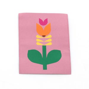 Könnte beinhalten: Ein rechteckiges, gewebtes Stoffetikett in zartem Rosa mit einer mittigen Grafik einer stilisierten Blume. Das Blumendesign besteht aus einem grünen Stiel mit Blättern, gelben horizontalen Blütenblättern, einer orangefarbenen Blüte und einem kleinen rosa Blütenblatt.