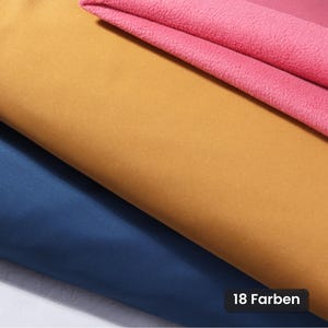 Peut inclure: Trois couches de tissu empilées. La couche supérieure est en polaire rose texturée, la couche intermédiaire est un tissu jaune moutarde lisse et la couche inférieure est un tissu bleu foncé lisse. Une étiquette noire affiche 18 Farben.