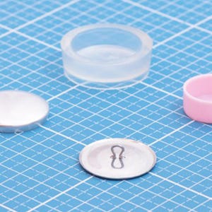 Può includere: Un bottone in metallo argentato con un occhiello in filo metallico sul retro, uno stampo in silicone traslucido e un piccolo contenitore circolare in plastica rosa. Questi oggetti sono disposti su un tappetino da taglio blu con griglia.