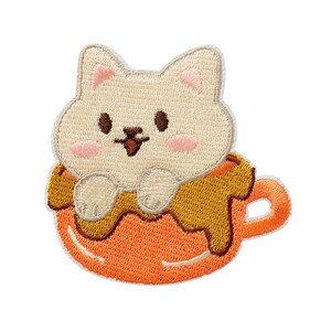 Pode incluir: Um emblema bordado apresentando um gato cor de creme com bochechas rosadas sentado dentro de uma xícara de chá laranja. O gato tem olhos e nariz castanho-escuros, com as patas dianteiras apoiadas na borda da xícara com líquido castanho.