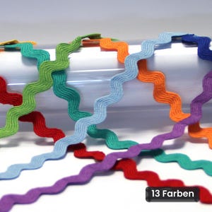 Op de afbeelding: Meerdere stroken golvend zigzagband in diverse kleuren, waaronder rood, groen, lichtblauw, oranje en paars. Het band heeft een consistent zigzagpatroon en wordt getoond tegen een witte achtergrond met de tekst 13 Farben.