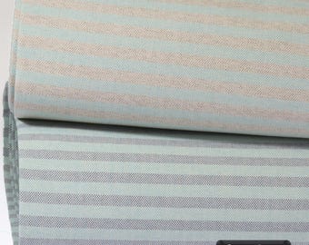 May include: Two folded pieces of fabric featuring horizontal stripes. The top piece displays alternating light blue and beige stripes, while the bottom piece shows alternating light blue and grey stripes. A black label reads 6 Farben.