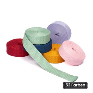 Peut inclure: Un ensemble de cinq rouleaux de sangle en tissu aux couleurs rouge, vert clair, bleu foncé, jaune, violet clair et rose. Un rouleau vert clair est partiellement déroulé. Une étiquette rectangulaire noire affiche le texte 52 Farben.
