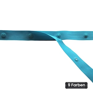 May include: A bright turquoise fabric tape featuring evenly spaced, integrated snap fasteners along its length. The tape is shown in a horizontal orientation with one section angled downward, and a small black label displays the text 9 Farben.