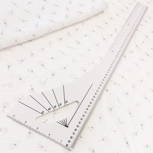 Op de afbeelding: Een witte stof met een herhalend patroon van kleine, metallic zilveren paardenbloemzaadjes. Een wit plastic kleermakersliniaal met zwarte markeringen en hoekaanduidingen ligt erbovenop en toont een lengtemeting tot 60 centimeter.