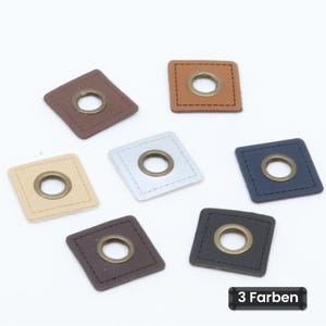Può includere: Una collezione di sette toppe quadrate in vari colori, tra cui beige, marrone, nero, bianco e blu navy. Ogni toppa presenta un occhiello metallico centrale color ottone e un bordo cucito. Il testo 3 Farben è visibile.