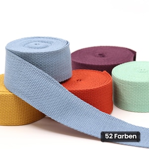 Pode incluir: Cinco rolos de fita de algodão tecida em várias cores sólidas, incluindo azul claro, amarelo mostarda, laranja escuro, roxo profundo e verde menta. Uma longa tira da fita azul claro estende-se para a frente com uma etiqueta preta 52 Farben.