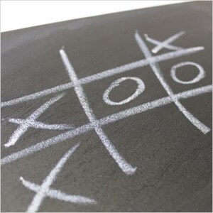 Könnte beinhalten: Eine dunkelgraue Tafeloberfläche mit einer weißen Kreidezeichnung eines Tic-Tac-Toe-Gitters. Das Spielfeld enthält zwei weiße Kreise und zwei weiße Kreuze, die mit Kreide in die Felder des Gitters gezeichnet wurden.