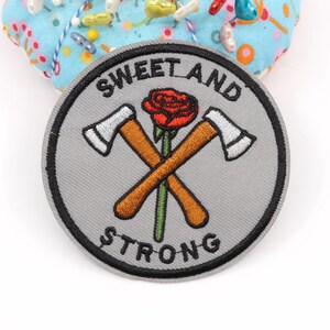 Pode incluir: Um emblema bordado circular com fundo cinza e borda preta. O design apresenta dois machados cruzados com cabos marrons e lâminas prateadas, centralizados atrás de uma rosa vermelha com caule verde. O texto SWEET AND STRONG está bordado em preto.