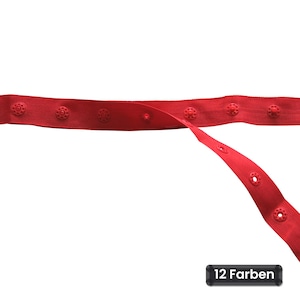 Könnte beinhalten: Ein rotes Stoffband mit einer Reihe von gleichmäßig verteilten Druckknöpfen entlang seiner Länge. Das Band ist horizontal ausgerichtet, wobei ein Teil nach unten verläuft, und ein kleines schwarzes rechteckiges Etikett zeigt den Text 12 Farben.