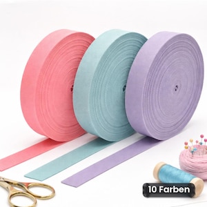Op de afbeelding: Drie grote rollen plat stoffen lint in roze, lichtblauw en lavendel. Elke rol heeft een lang uiteinde van het lint dat naar voren uitsteekt. Een goudkleurige schaar, een kleine klos blauw garen en een speldenkussen met spelden zijn zichtbaar.