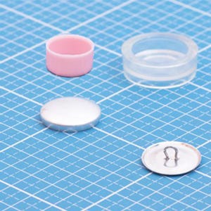 May include: A set of button-making components including a small pink cylindrical plastic mold, a clear circular plastic ring, a smooth silver-colored metal button cap, and a silver-colored metal button back featuring a central wire loop attachment.