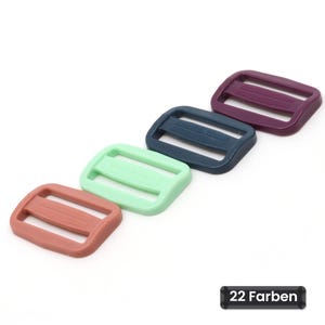 Könnte beinhalten: Vier rechteckige Kunststoff-Gurtversteller, die diagonal angeordnet sind. Die Farben sind gedecktes Rosa, helles Mintgrün, dunkles Marineblau und tiefes Pflaumenviolett. Jeder Versteller hat einen Mittelsteg mit horizontalen Linien.