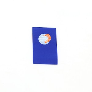 Può includere: Un'etichetta rettangolare in tessuto blu con una grafica circolare azzurra raffigurante un gamberetto arancione e il testo bianco "YOU'RE SHRIMP-LY THE BEST" stampato al centro del cerchio, progettata per essere applicata sui capi.