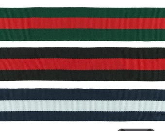 May include: Three strips of woven ribbon with a ribbed texture. The top strip is dark green with a central red stripe. The middle strip is black with a central red stripe. The bottom strip is navy blue with a central white stripe. Text reads 7 Farben.