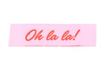 May include: A rectangular fabric clothing label in a light pink colour featuring the text Oh la la! embroidered or printed in a bold red script font. The label has a clean, straight-cut edge and a smooth, woven texture throughout the surface.