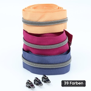 Könnte beinhalten: Ein Stapel aus drei aufgerollten Reißverschlüssen in Pfirsich, Magenta und Marineblau, jeweils mit dunklen Metallzähnen. Drei separate schwarze Reißverschlussschieber liegen vor dem Stapel und ein schwarzes rechteckiges Etikett zeigt den Text 39 Farben.