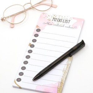 Op de afbeelding: Een rechthoekig to-do-lijstblok met een roze en gouden aquarelontwerp, voorzien van de tekst "TO DO LIST" en "Aber erstmal nähen!!" met een kolom van gedrukte grijze knopen, vergezeld door een zwarte balpen en een bril met gouden montuur.
