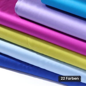 Può includere: Una pila di tessuto di raso piegato in vari colori, tra cui magenta, viola chiaro, blu brillante, giallo e azzurro. Il tessuto ha una superficie liscia e riflettente. Un'etichetta rettangolare nera nell'angolo riporta il testo 22 Farben.