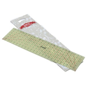 May include: A transparent yellow-tinted rectangular sewing ruler with a grid of red crosshairs and black measurement markings up to 20 centimeters. The ruler is attached to a light gray cardboard backing featuring white sewing icons and text.