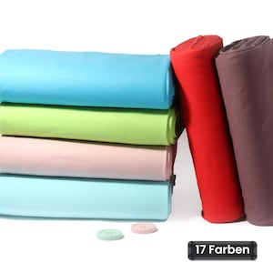 Könnte beinhalten: Ein Stapel aus vier gefalteten Stoffballen in Hellblau, Limettengrün, Blassrosa und Cyan sowie zwei aufrecht stehende Rollen in Rot und Dunkelbraun. Zwei kleine runde Knöpfe in Mint und Rosa liegen davor, mit einem schwarzen Etikett 17 Farben.