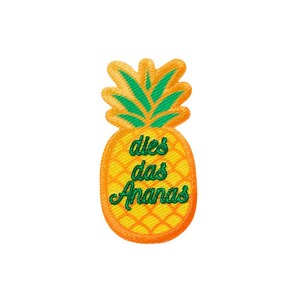 Op de afbeelding: Een geborduurde opstrijkbare patch in de vorm van een ananas met een geel lichaam en groene bladeren. De ananas heeft een ruitvormige textuur en de groene geborduurde tekst "dies das Ananas" staat gecentreerd op de voorkant van de vrucht.
