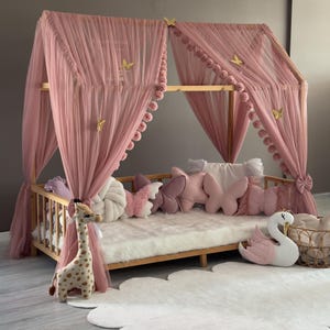 Puede incluir: Una cama con dosel de madera con cortinas de tul rosa y adornos de mariposas. La cama está amueblada con almohadas con forma de mariposa, un peluche de jirafa y una almohada con forma de cisne. La cama está sobre una alfombra blanca y esponjosa.