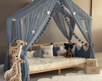 May include: A wooden bed frame with a blue canopy, adorned with star-shaped decorations and pom-poms. The bed is furnished with pillows, a white faux fur blanket, and two giraffe plush toys. Two small cardboard boxes are on the side.