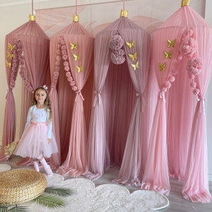 May include: Four pink tulle canopy beds with gold butterfly and pom-pom accents. A young girl is standing in front of the canopies wearing a pink tulle skirt and a white long-sleeved shirt.