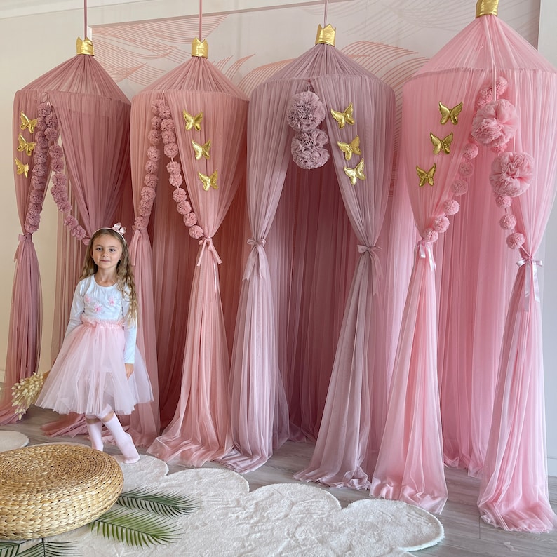 May include: Four pink tulle canopy beds with gold butterfly and pom-pom accents. A young girl is standing in front of the canopies wearing a pink tulle skirt and a white long-sleeved shirt.