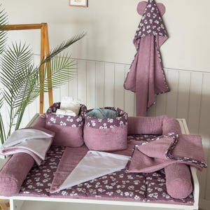 Peut inclure: Ensemble de table à langer pour bébé dans les tons mauve et blanc. L'ensemble comprend un matelas à langer, deux paniers de rangement, une serviette à capuche avec des oreilles d'ours et plusieurs petites serviettes. Les articles sont décorés d'un motif floral.