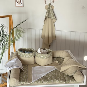 May include: A baby changing station set with a floral print in green and white. The set includes a changing pad, two storage baskets, a hooded towel, and a bolster pillow. The items are in neutral tones of beige and white.