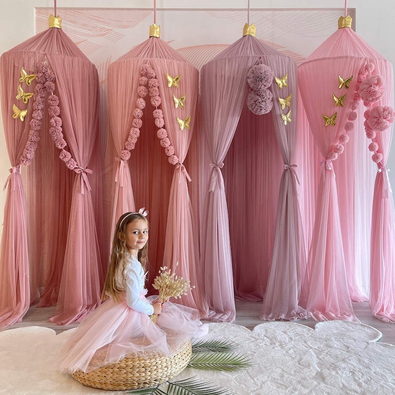 May include: Four pink and mauve tulle canopy beds with gold butterfly decorations and pom pom trim. A young girl sits in front of the canopies wearing a pink tulle skirt and a white shirt.