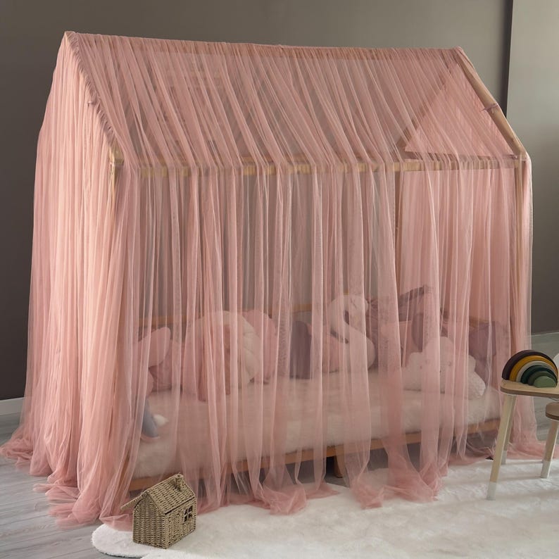May include: A pink canopy bed with a wooden frame and sheer, flowing fabric. The bed is decorated with pillows and a small decorative house. The bed is on a white rug.