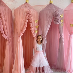 May include: A young girl stands in a pink and mauve canopy bed with a white tulle skirt and a white shirt. The canopy is decorated with pom poms and gold butterflies.