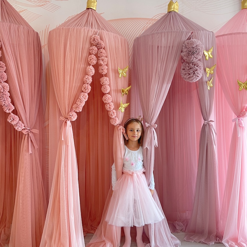 May include: A young girl stands in a pink and mauve canopy bed with a white tulle skirt and a white shirt. The canopy is decorated with pom poms and gold butterflies.