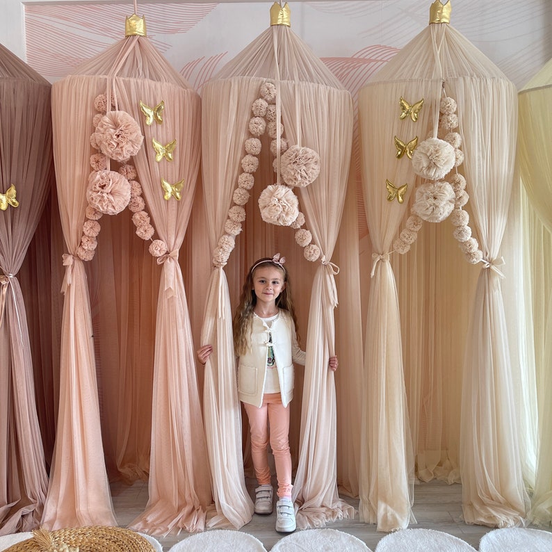 Puede incluir: Una ni&ntilde;a peque&ntilde;a se encuentra frente a tres camas con dosel de color melocot&oacute;n transparente con detalles de mariposas doradas y ribetes de pomp&oacute;n. Los doseles est&aacute;n hechos de una tela suave y fluida y crean un ambiente caprichoso y acogedor.