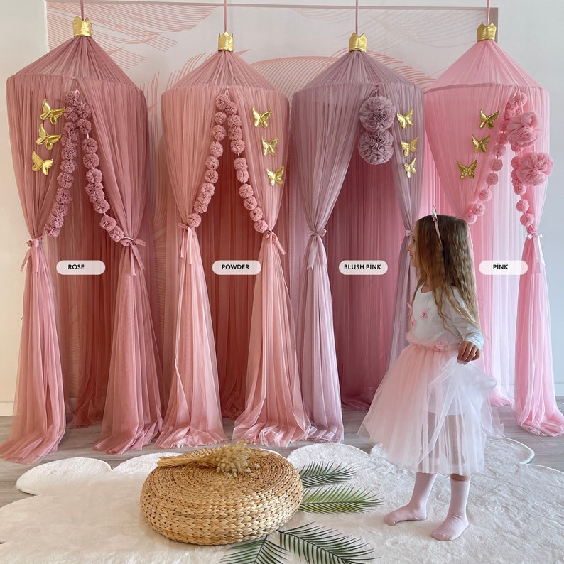 May include: Four pink and blush colored tulle canopy beds with gold pom poms and butterflies. The canopies are labeled "Rose", "Powder", "Blush Pink", and "Pink". A young girl wearing a white shirt and a pink tulle skirt stands in front of the canopies.