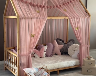 May include: A wooden house-shaped bed frame with pink tulle curtains and decorative butterflies. The bed is adorned with various pillows in shades of pink and gray, and a fluffy white rug. A gold wire basket and two beige boxes are in the foreground.