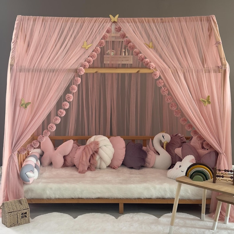 May include: A pink canopy bed with sheer curtains, adorned with butterfly decorations and pom-pom garlands. The bed is furnished with various pillows, including butterfly and swan-shaped ones, and sits on a fluffy white rug.