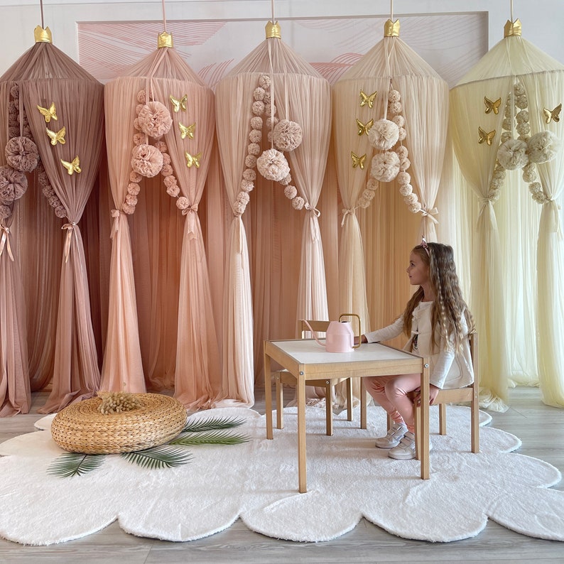 Puede incluir: Cinco cortinas de dosel de tela transparente con ribete de pompones y detalles de mariposas doradas. Un ni&ntilde;o se sienta en una mesa peque&ntilde;a de madera y sillas frente a las cortinas.