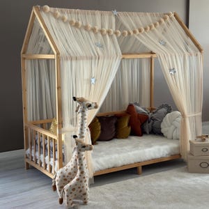 Puede incluir: Una cama de madera con forma de casa con un colchón de piel sintética blanca y cortinas transparentes. Cojines decorativos de varios colores y dos jirafas de peluche están frente a la cama. La cama está decorada con adornos en forma de estrella y una guirnalda de pompones.