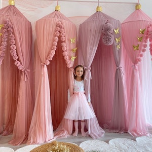 May include: Three pink tulle canopy beds with gold butterfly decorations and pom poms. A young girl wearing a white shirt and a pink tulle skirt stands in front of the canopies.