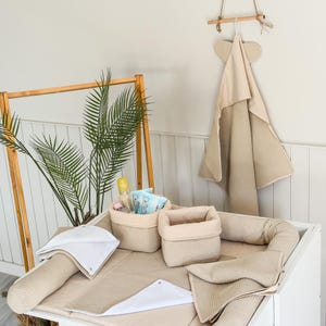 Peut inclure: Un ensemble de change pour bébé aux tons neutres. Il comprend un matelas à langer, un coussin, deux paniers de rangement et une serviette à capuche. Les articles sont beiges et blancs, créant une ambiance apaisante pour la chambre de bébé.