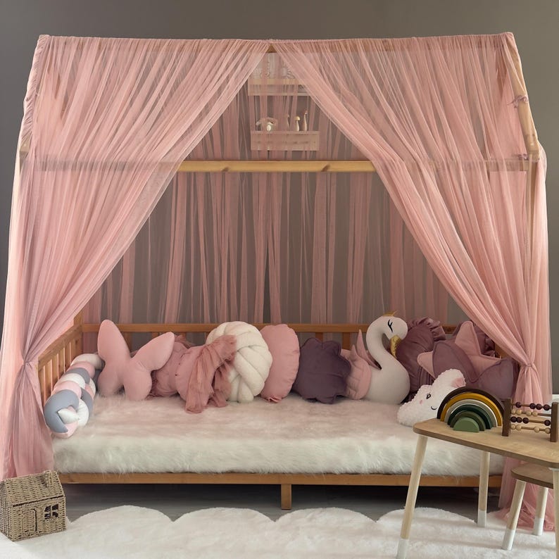 May include: A wooden bed frame with a pink canopy and decorative pillows. The bed is adorned with a white faux fur rug and a variety of plush pillows in shades of pink, purple, and white. A small wooden table with toys sits beside the bed.