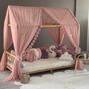 May include: A wooden canopy bed with pink tulle drapes and decorative features. The bed has a fluffy white rug, decorative pillows, and a small house-shaped basket. Gold butterfly and pom-pom details enhance the whimsical design.
